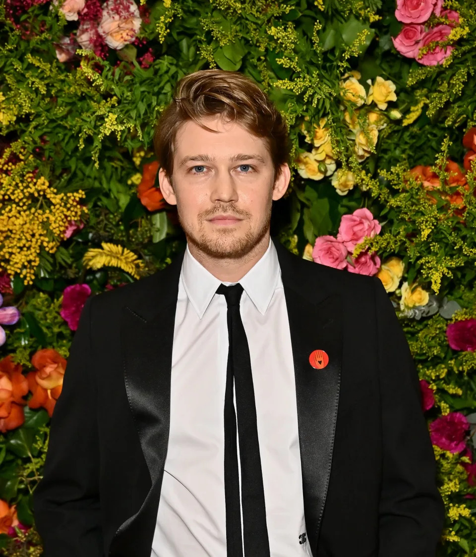 Joe Alwyn standing in front of a floral backdrop wearing a black suit with a white shirt and black tie