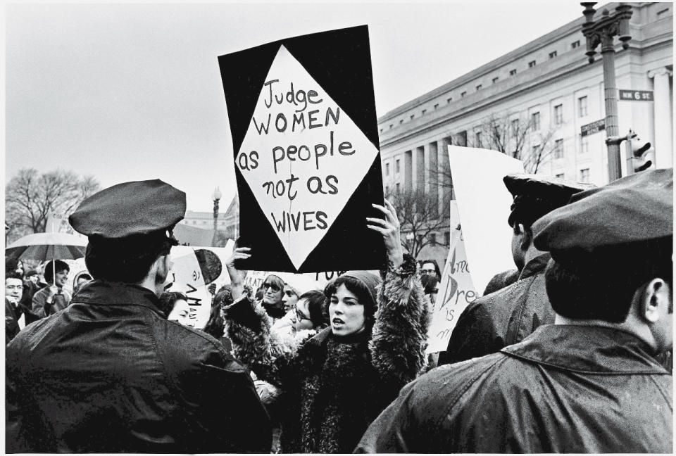 A young American woman holds up a sign as she protests for women's rights in front of the Federal Trade Commission headquarters while policemen look on during Richard Nixon's inauguration weekend, Washington, DC, January 18-21, 1969. Her sign reads 'Judge women as people not as wives.'&nbsp;