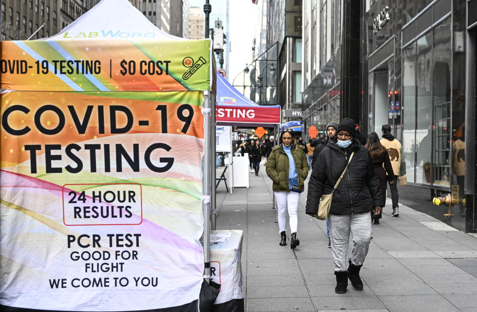 Pedestrians, one of whom wears a face mask, walk by a COVID-19 testing site on a sidewalk.