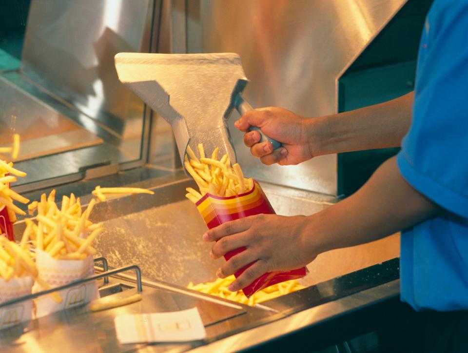 A worker at McDonald's fills a red and yellow fries container with freshly cooked French fries using a metal scoop