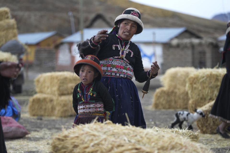 Standing with her daughter, a woman weaves next to fodder brought to feed the starving animals of the Cconchaccota community, in the Apurimac region of Peru, Friday, Nov. 25, 2022. The fodder is the long-delayed drought response from the regional authorities to feed for the surviving sheep, cattle, alpacas and llamas. (AP Photo/Guadalupe Pardo)