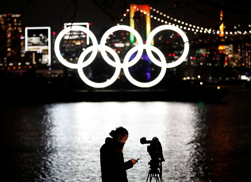 A media member reports while the giant Olympic rings are illuminated after being reinstalled at Odaiba Marine Park, amid the coronavirus disease (COVID 19) outbreak, in Tokyo
