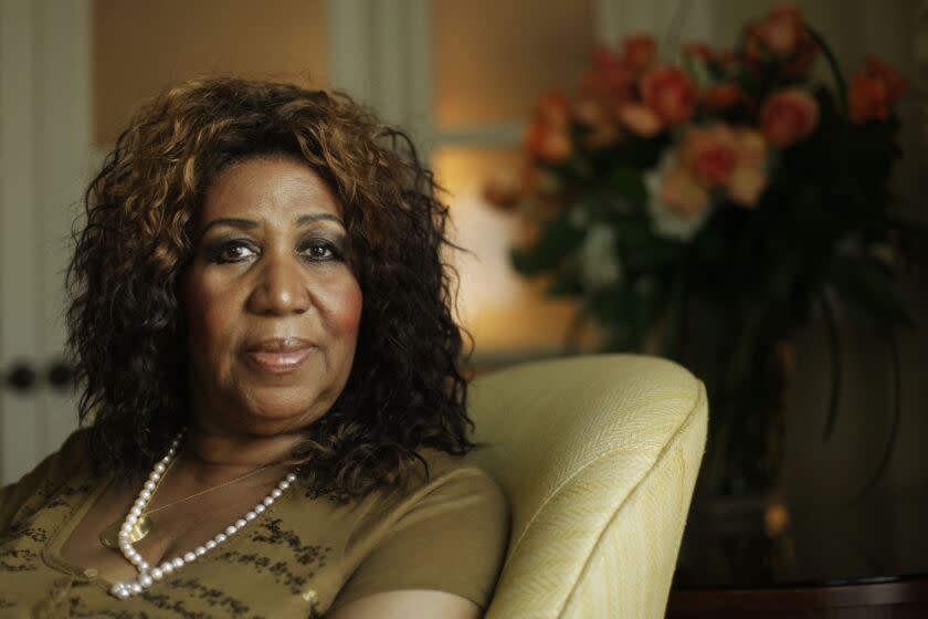 Aretha Franklin is sitting in a beige couch wearing a brown shirt and pearl necklace, posing with a serious face