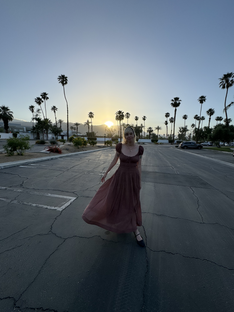 Person in a full-length dress standing in an empty parking lot with palm trees and sunset in the background
