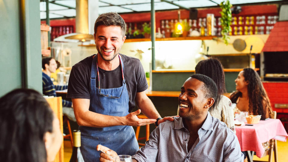 Couple sitting opposite each other in small restaurant, waiter standing and socialising, relaxed and friendly, customer service.