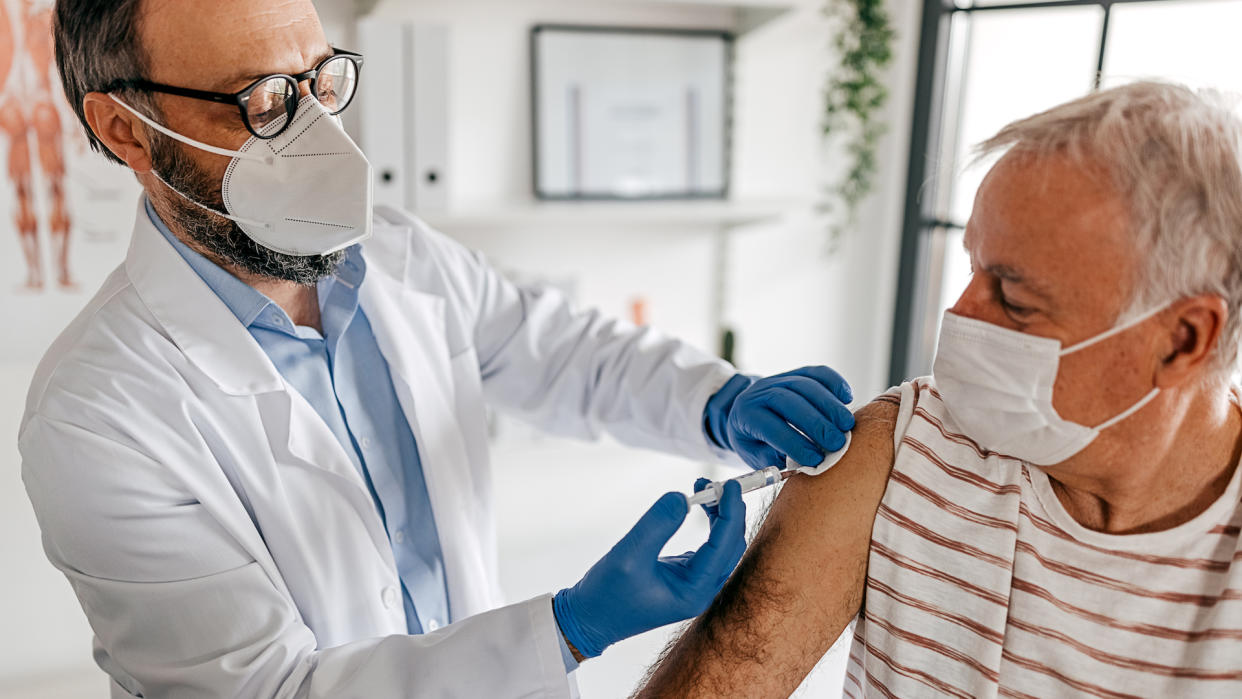  A male doctor wearing a white N95 mask gives a vaccine to an older male patient wearing a white surgical mask. 