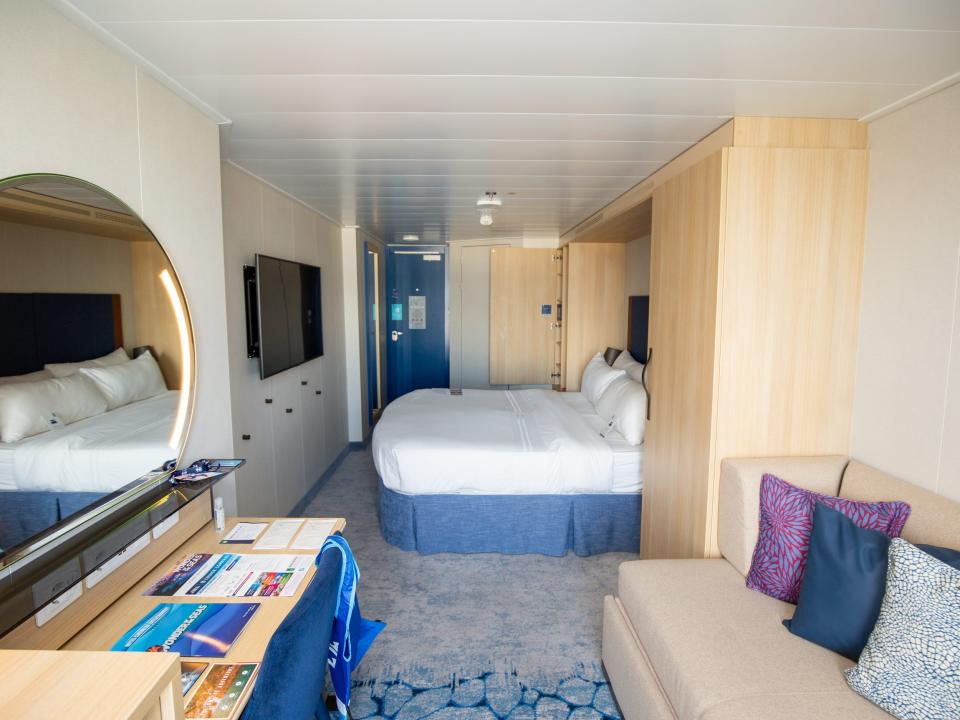 A stateroom with a double bed, a cream-colored couch, and a large mirror next to a TV.