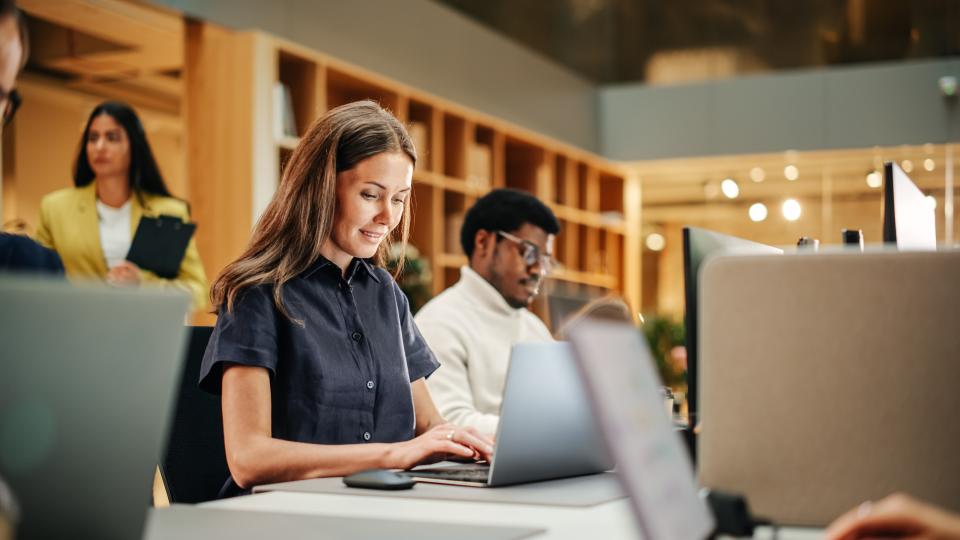 A woman working on a laptop in a shared working space sitting next to a man working at a computer