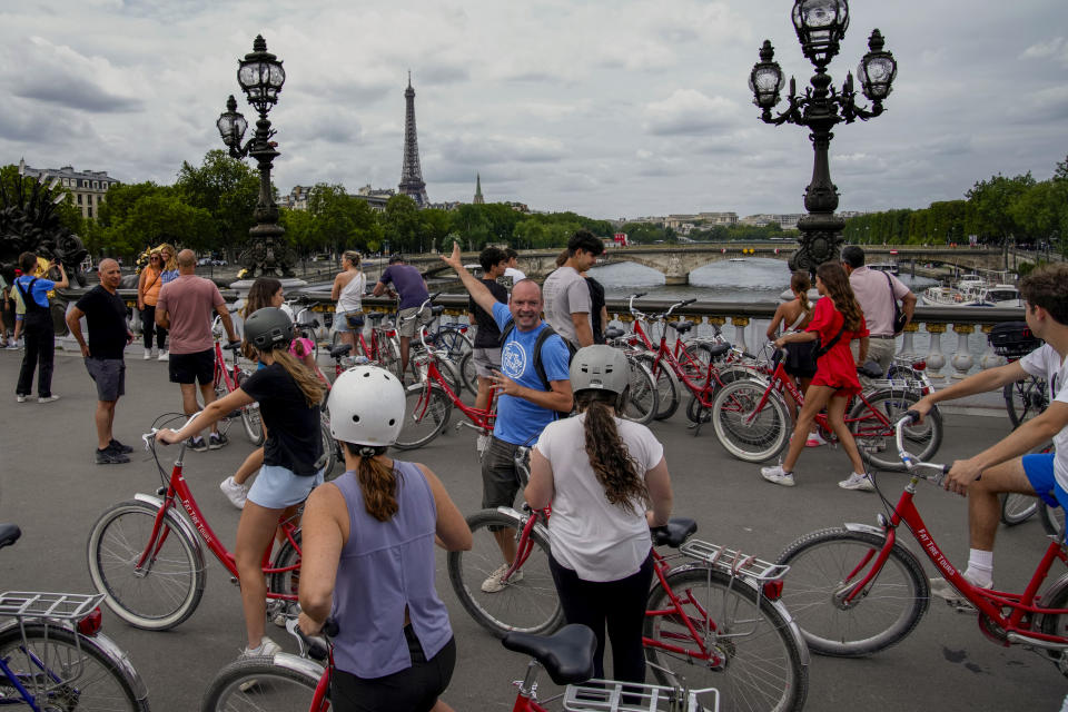 Tourists on a bicycle tour watch the Eiffel Tower during a stop at a bridge atop the Seine River in Paris, Tuesday, July 4, 2023. Crowds are packing the Colosseum, the Louvre, the Acropolis and other major attractions as tourism exceeds 2019 records in some of Europe’s most popular destinations. While European tourists helped the industry on the road to recovery last year, the upswing this summer is led largely by Americans, who are lifted by a strong dollar and in some cases pandemic savings. (AP Photo/Christophe Ena)