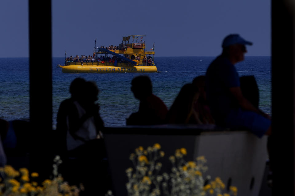 Tourist in silhouettes sit at a kiosk as a cruise boat with tourist enjoy the sea at Limanaki beach in southern coastal resort of Ayia Napa in southeast Mediterranean island of Cyprus, Sunday, June 5, 2022. Hundreds of Russian and Ukrainian Orthodox faithful visiting Cyprus would stream daily past the icon of the Virgin Mary at Kykkos Monastery to venerate the relic that tradition dictates was fashioned by Luke the Evangelist and blessed by the Virgin herself. But a European Union ban on flights to and from Russia as a result of Russia's invasion of Ukraine has meant a loss of 800,000 vacationers - a fifth of all tourists to Cyprus in record-setting 2019. (AP Photo/Petros Karadjias)