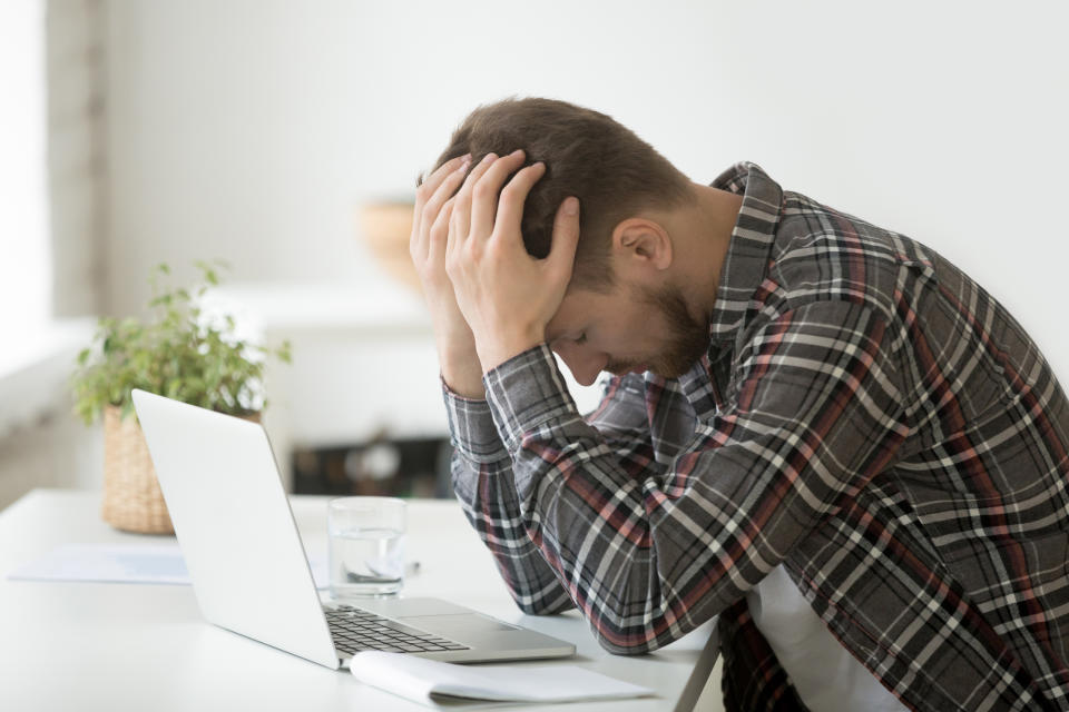 A man in a plaid shirt puts his head in his hands in anguish sitting in front of his laptop.