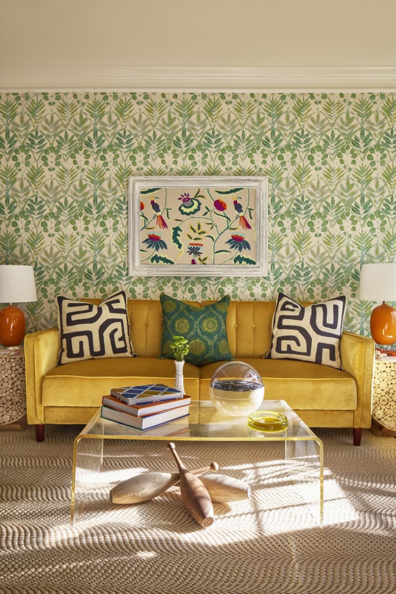 green floral patten wallpaper, yellow couch with patterned and colorful pillows, glass coffee table with gold trim