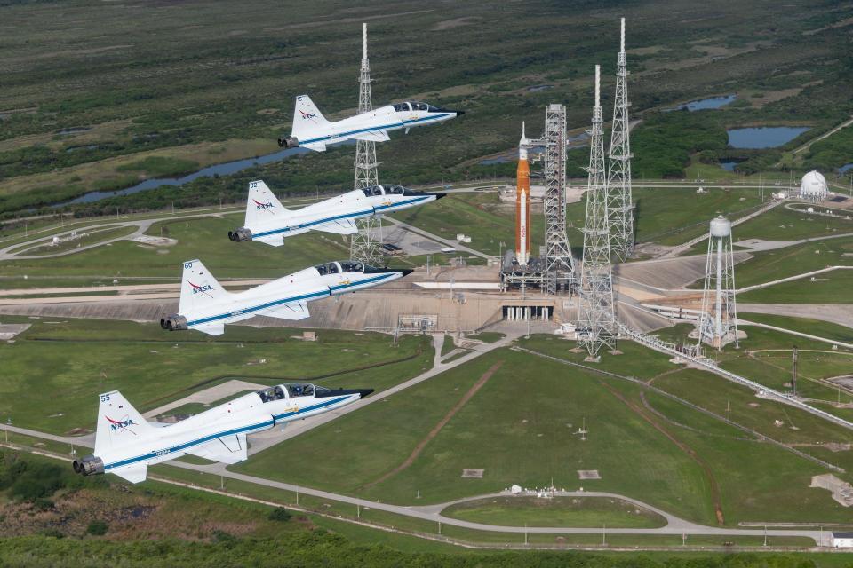 four white jets are flying above a large orange rocket standing on a launch pad