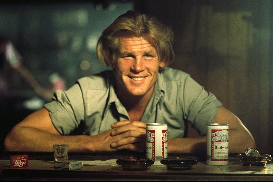 Nick Nolte smiling with hands on a bar table next to beer cans
