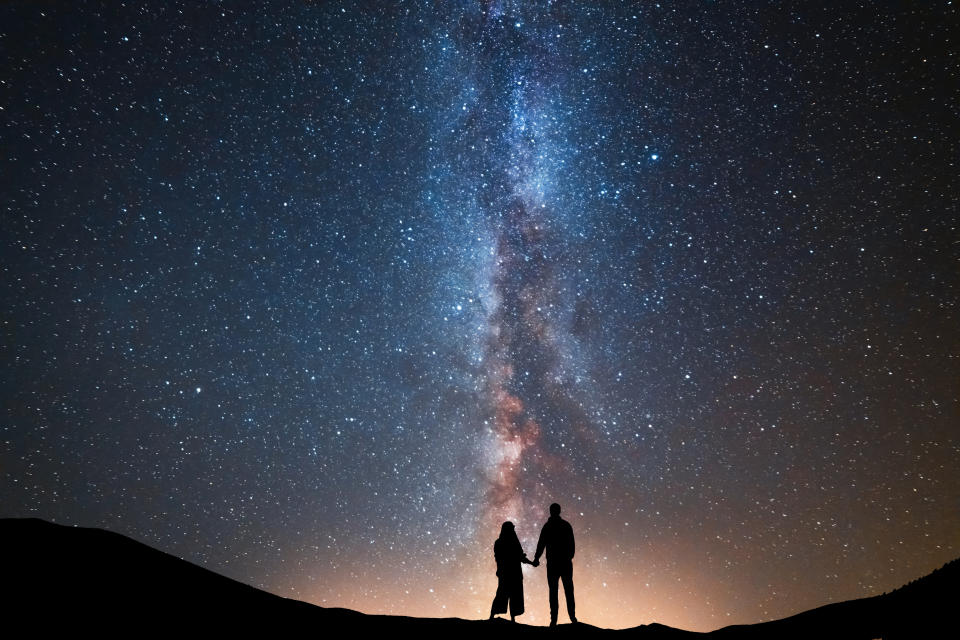 Two people holding hands, silhouetted against a star-filled sky and the Milky Way