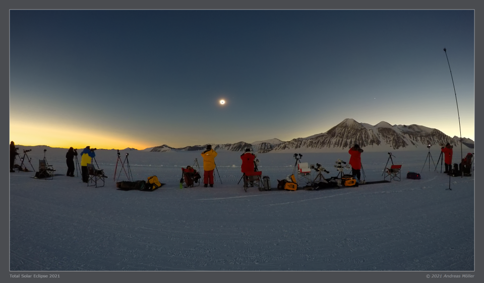 A row of photographers stands in the snow, with mountains in the distance to the right.  The darkening sky contains a small black dot surrounded by bright light.