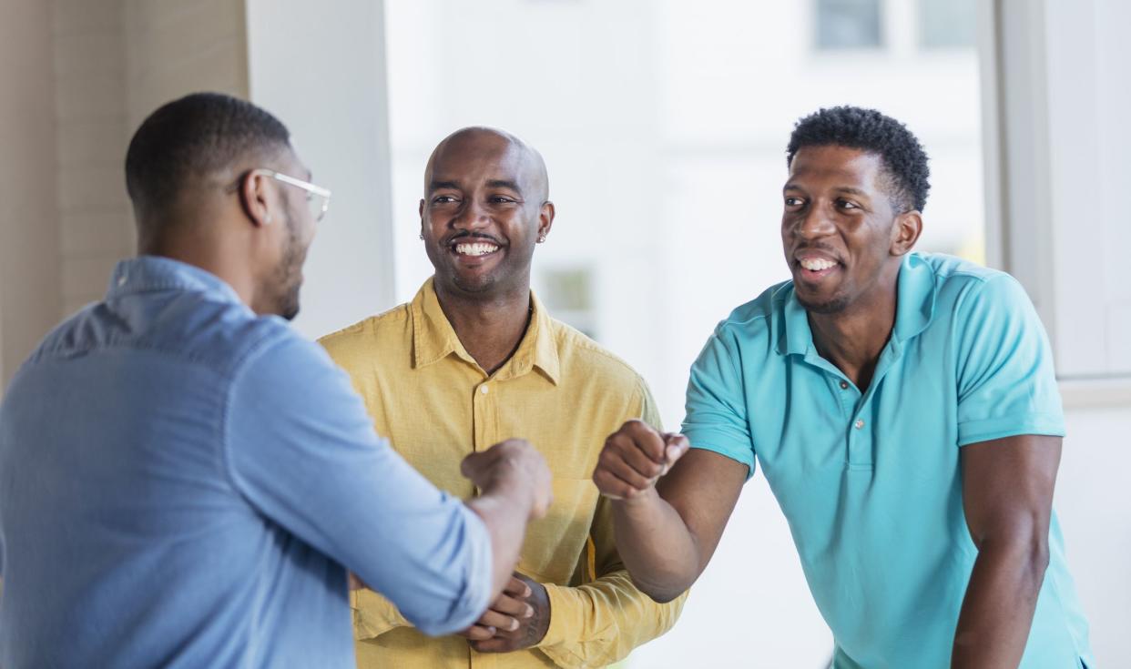 A group of three African-American men standing face to face indoors, smiling and conversing. Two of them are bumping fists. They are mid adult men in their 30s.