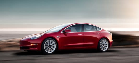 A red Tesla Model 3, a sleek compact luxury sedan, on a coastal road at sunset.