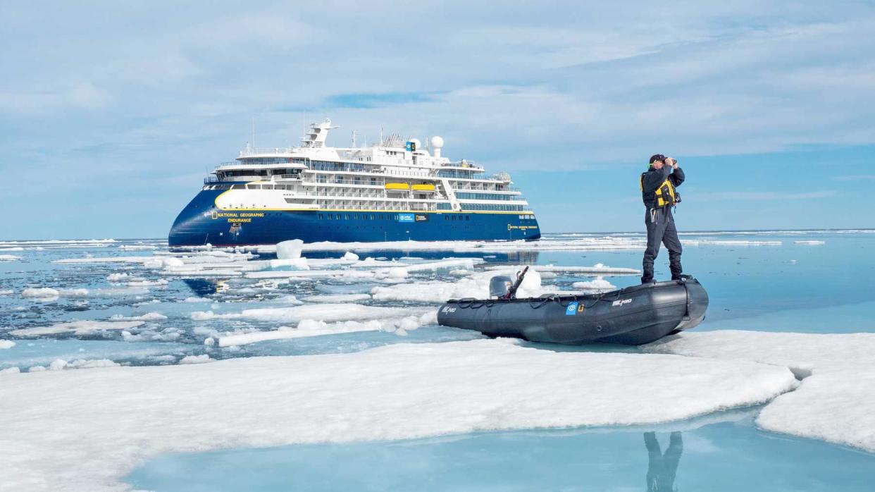 A man stands on a zodiac in Greenland, with the National Geographic Endurance expedition ship in the background