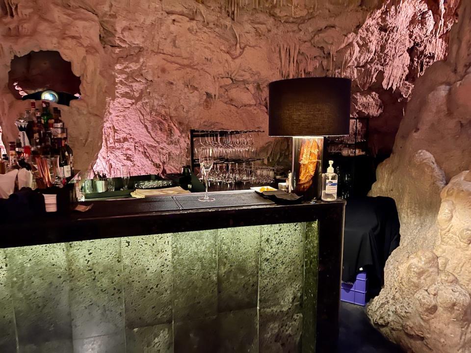 A bar counter inside a restaurant that's in a cave. There are bottles of alcohol stacked on the shelf on the side, and a row of wine glasses behind the counter.