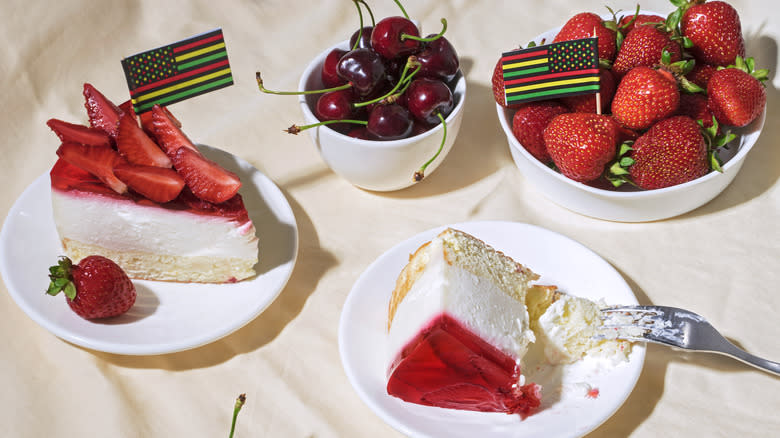 Plated strawberry cheesecake next to bowls of cherries and strawberries served with small Juneteenth flags