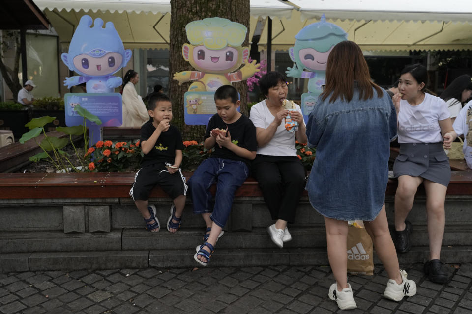 Tourists eat snacks near cutouts of official mascots of the 19th Asian Games in Hangzhou in eastern China's Zhejiang province, on June 26, 2023. The Asian Games open in two weeks, the first multi-sport international event in China since pandemic restrictions were lifted there about nine months ago. Unmatched for size, the Asian Games may even surpass the Olympics for controversy, power politics and intrigue. (AP Photo/Ng Han Guan, File)