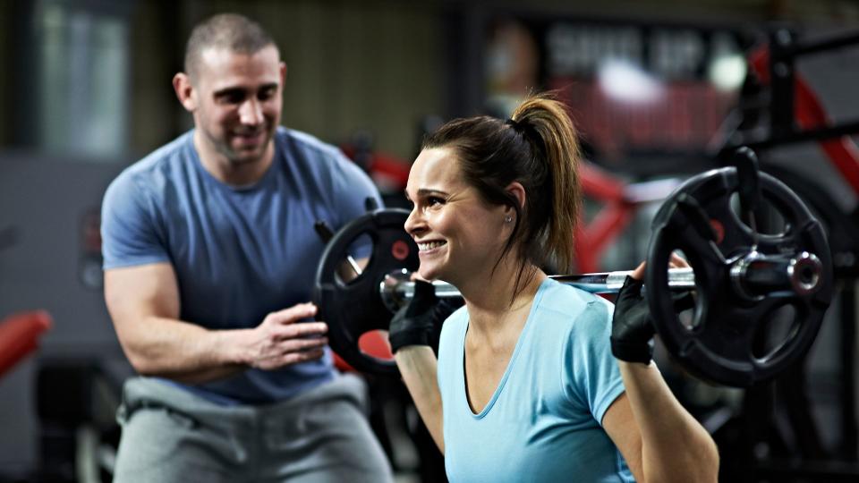 Woman moving down into a squat with weighted barbell, personal trainer helping on one side