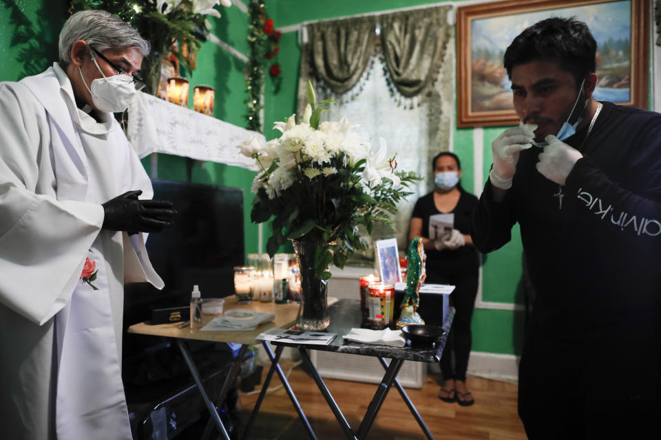 Family members receive communion by their own hand as the Rev. Fabian Arias, left, performs an in-home service beside the remains of Raul Luis Lopez who died from COVID-19 the previous month, Saturday, May 9, 2020, in the Corona neighborhood of the Queens borough of New York. (AP Photo/John Minchillo)