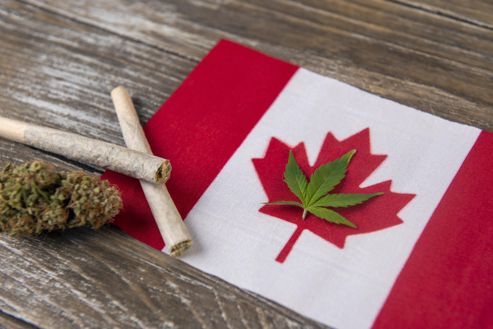 A cannabis leaf lying with the outline of the Canadian flag's red maple leaf, with rolled joints and a cannabis bud next to the flag.