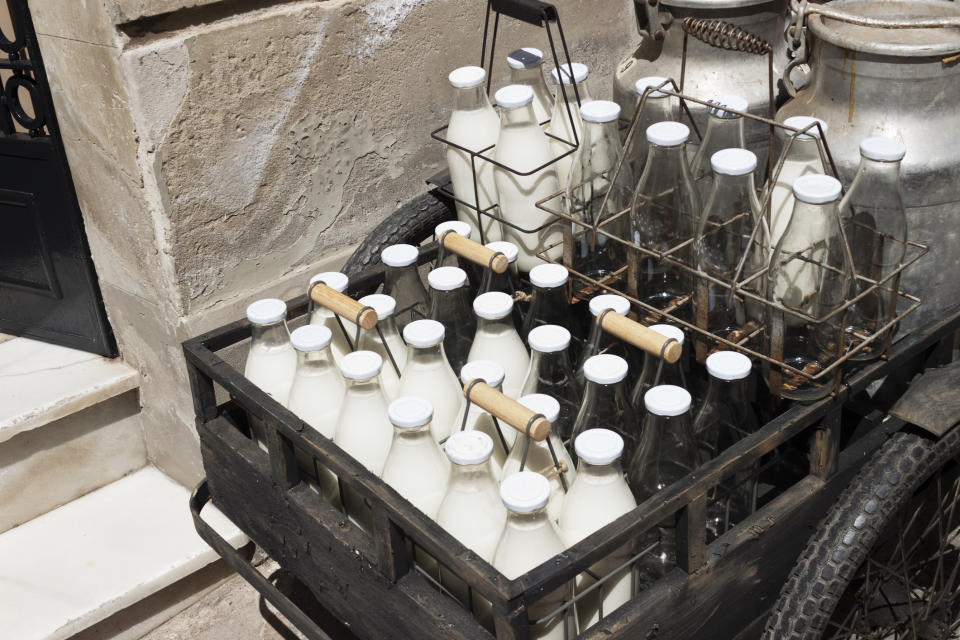 An old-fashioned wooden cart is filled with multiple glass milk bottles, some in crates, alongside metal milk containers, outside a stone building