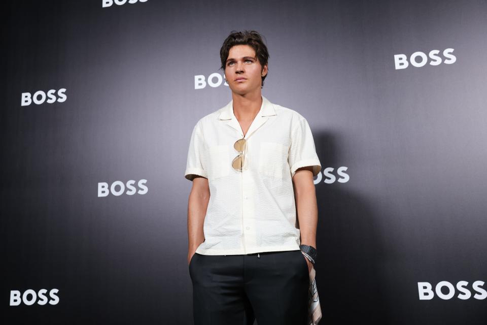 will peltz wearing a white button down, black pants, and sunglasses hanging on his shirt, in front of a black background branded with Boss for Hugo Boss
