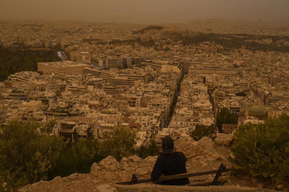 A tourist sits on a bench at the Lycabettus hill as the city of Athens with the ancient Acropolis hill is seen at the background, on Tuesday, April 23, 2024. The Acropolis and other Athens landmarks took on Martian hues Tuesday as stifling dust clouds blown across the Mediterranean Sea from North Africa engulfed the Greek capital. (AP Photo/Petros Giannakouris)