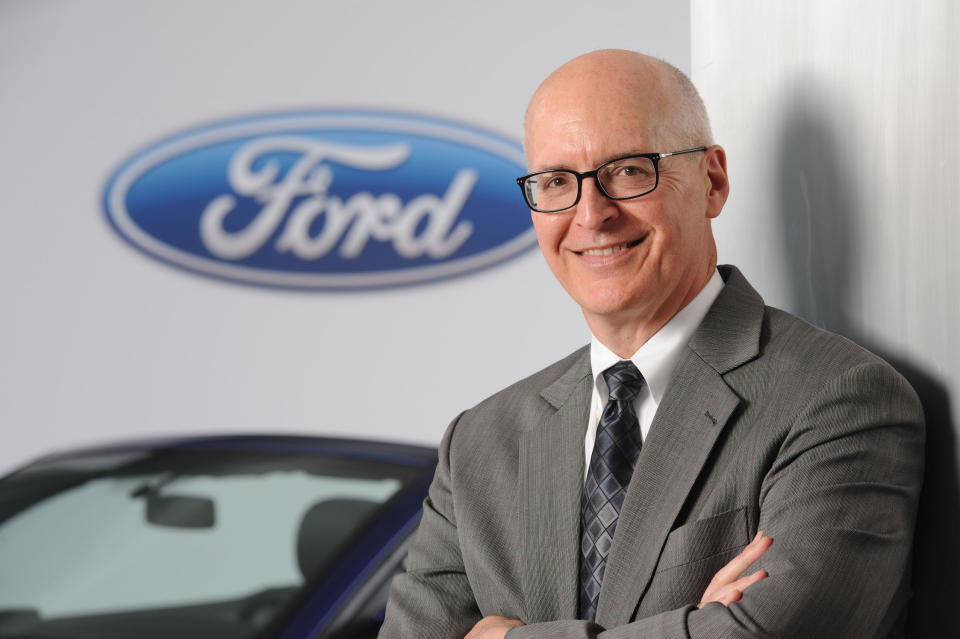 Shanks is shown standing in front of a blue Ford Mustang, and a Ford logo on a white backdrop behind the car.