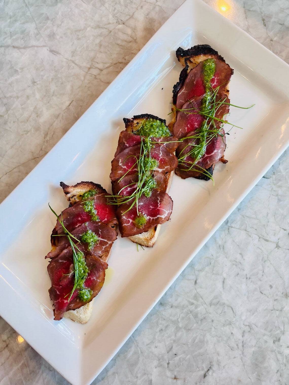 Panacea’s smoked carpaccio appetizer with black garlic, sliced baguette and herb pistou.