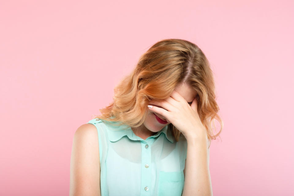 Woman against pink background covering her lowered face in apparent exasperation.
