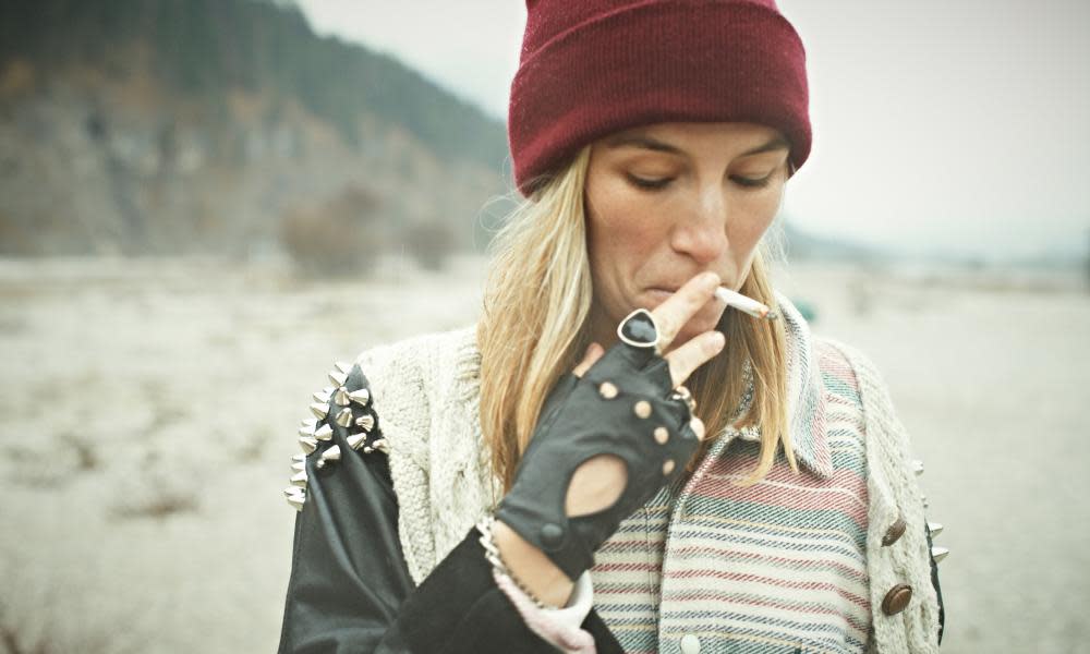 Woman in red woollen hat with long blond hair smoking roll-up cigarette on beach with cliffs in background