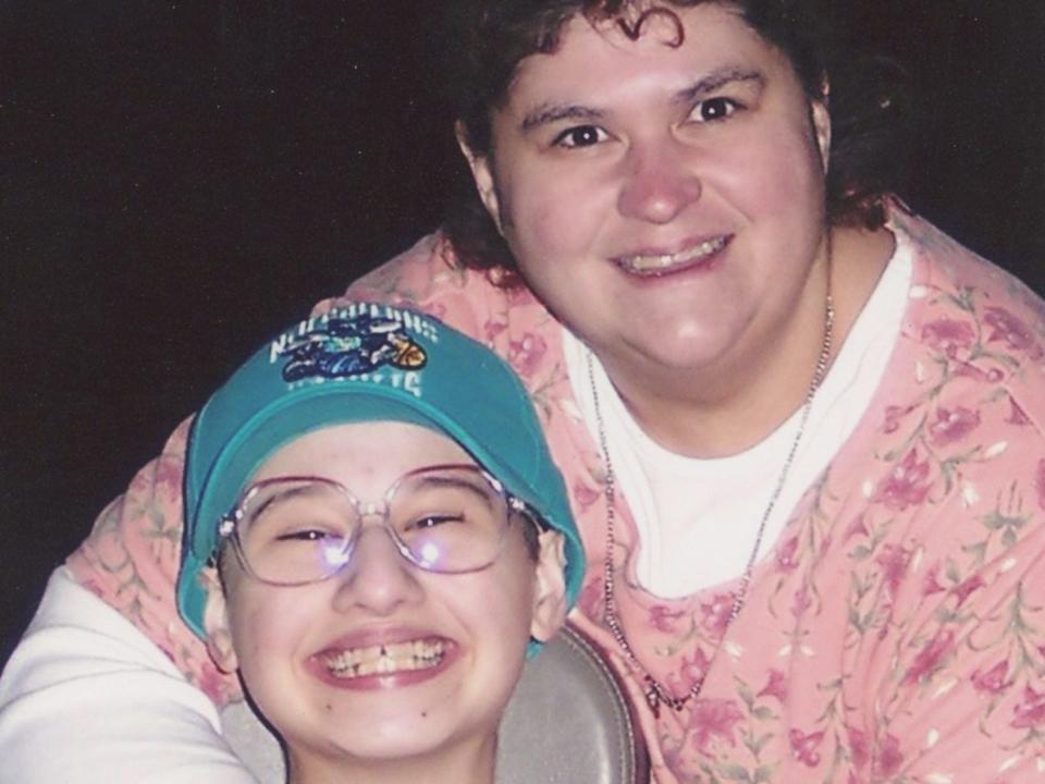 dee dee blanchard holding her arms around gypsy rose blanchard, who is smiling widely, sitting in a chair, and wearing a hat on her head