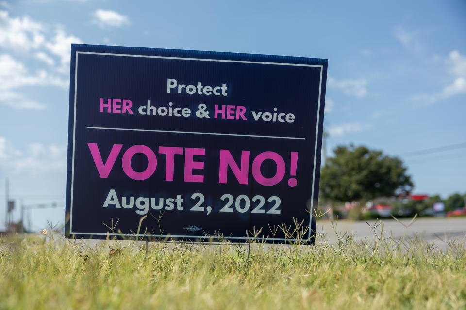 A sign in pink lettering on a black background standing in a field says: Protect HER choice & HER voice; VOTE NO! August 2, 2022.