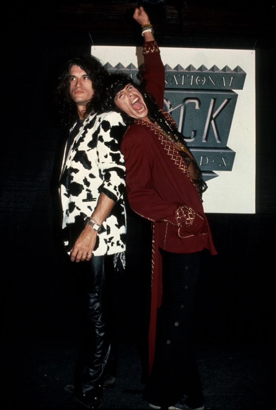 joe perry and steven tyler stand back to back in front of a poster, perry is wearing a cow print suit jacket and black pants, tyler is wearing a red jacket with gold accents and has one arm thrust over his head in the arm