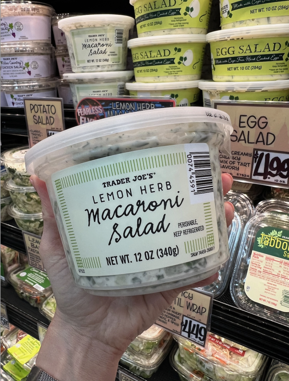 Hand holding a container of Trader Joe's Lemon Herb Macaroni Salad in front of a display of other prepared salads