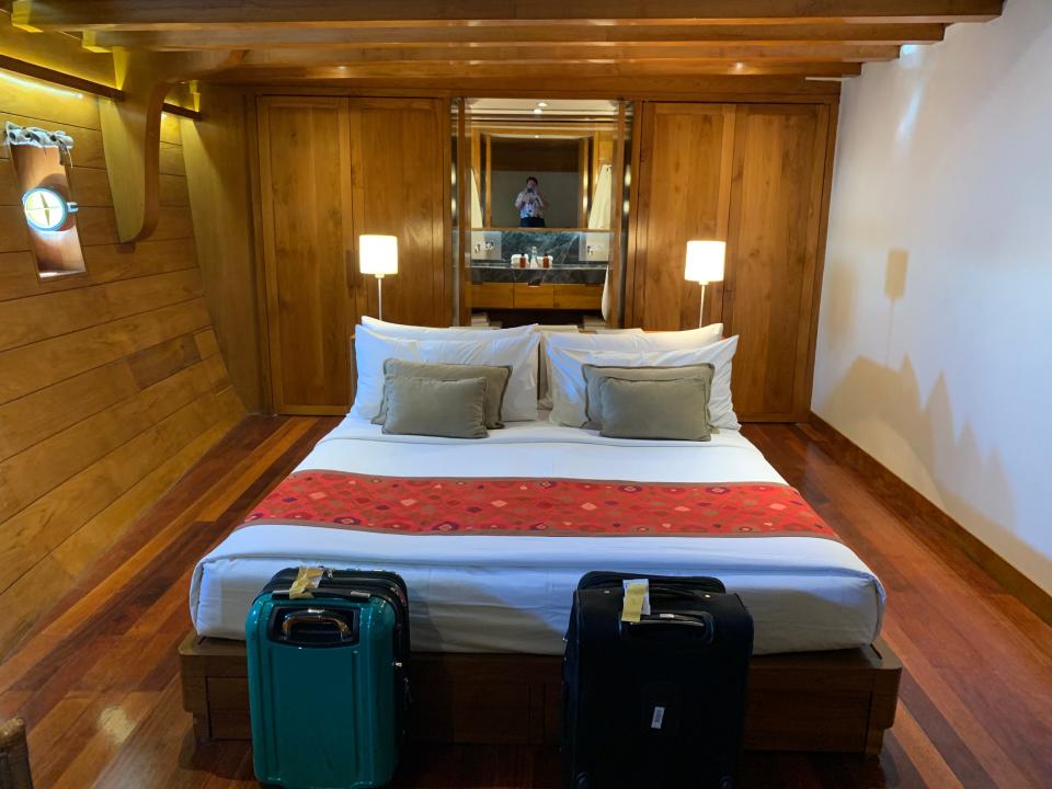 A ship cabin with wood walls, luggage in front of a bed, lamps, and a bathroom.