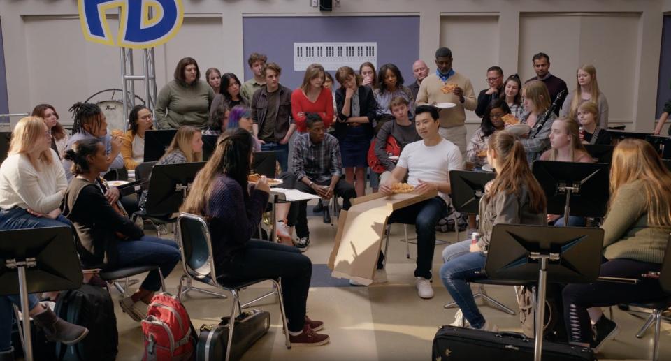 A class of students listens to a man talk while eating pizza.