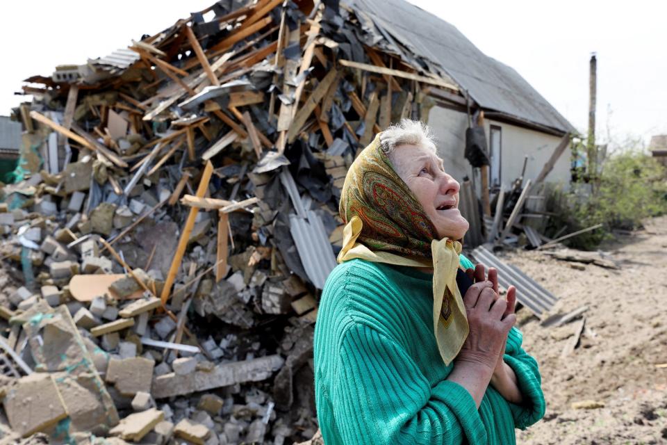 A woman wearing a headscarf and a green knitted top looks to the sky with a pained expression in front of a destroyed house