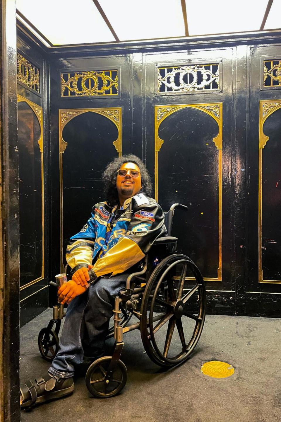 Saptarshi Majumdar wearing a racing jacket sits in a wheelchair in an elevator with black walls and gold trim.