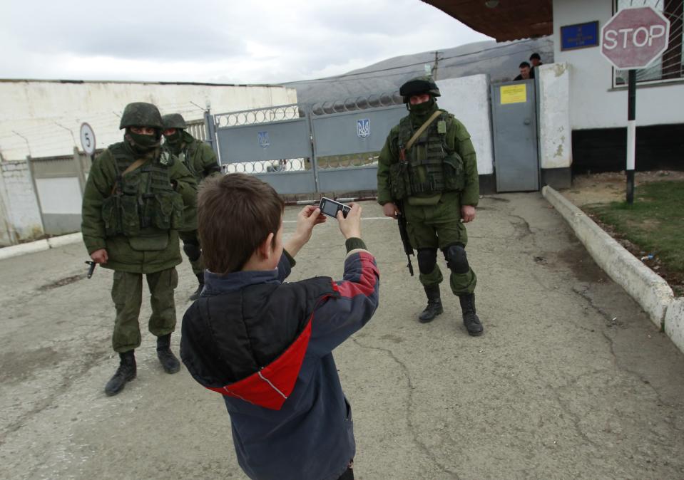A boy photographs military personnel, believed to be Russian servicemen, standing outside the territory of a Ukrainian military unit in the village of Perevalnoye outside Simferopol