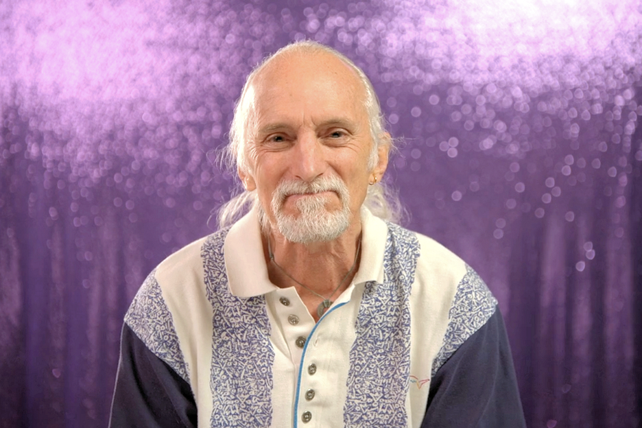 an older gentleman with a ponytail sits in front of a glittery backdrop. he smiles with his mouth closed in the shape of a line