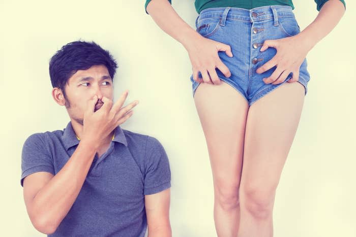 A person holds their nose while looking at another person's shorts. The person with shorts stands with their hands on their thighs