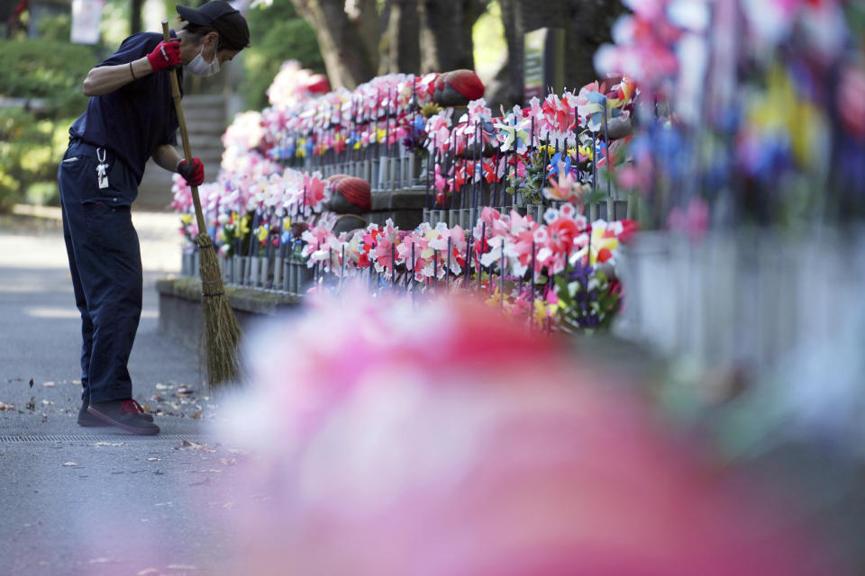 An employee wearing a protective mask to help curb the spread of the coronavirus sweeps near pinwheels whirl in the breeze at rows of small stone statues of "jizo" representing the unborn children at a temple in Tokyo Thursday, Oct. 29, 2020. The Japanese capital confirmed more than 220 new coronavirus cases on Thursday. (AP Photo/Eugene Hoshiko)