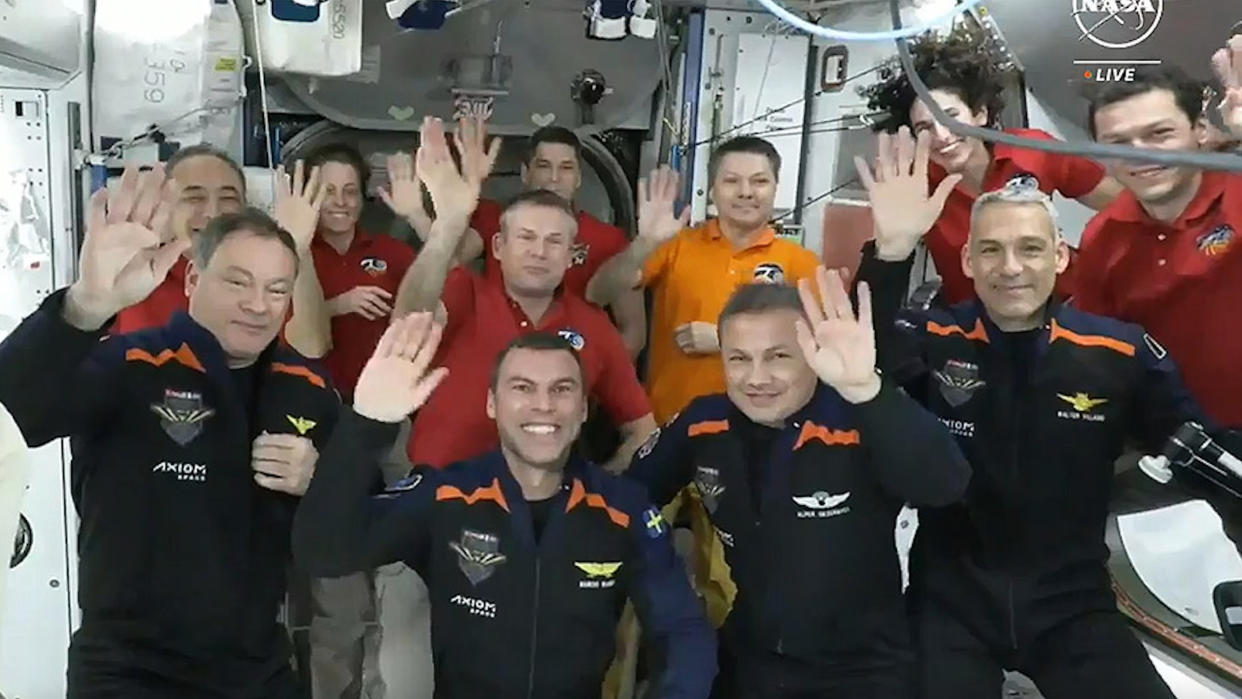  11 people in flight suits wave to the camera aboard the white-walled international space station. 