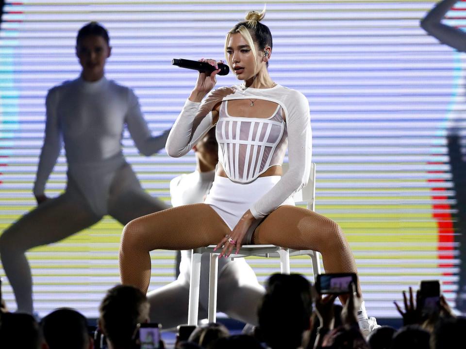 Dua Lipa sits in a chair on a stage in white shorts and a white see-through top.